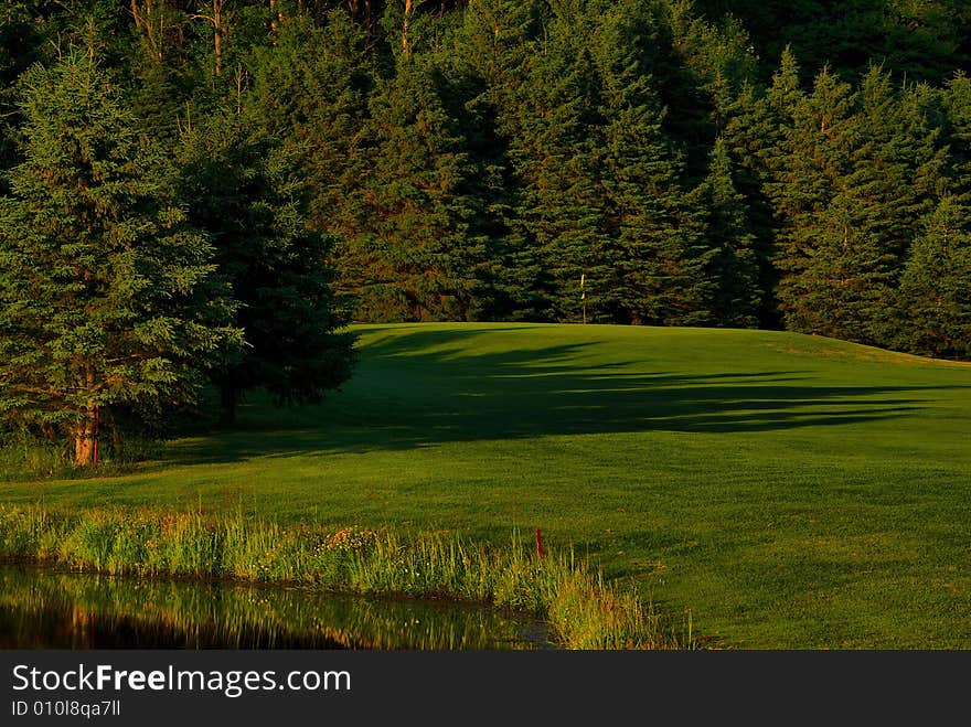 A short shot over the water to an elevated green.  The sun is casting long shadows across the fairway during a late afternoon round of golf.