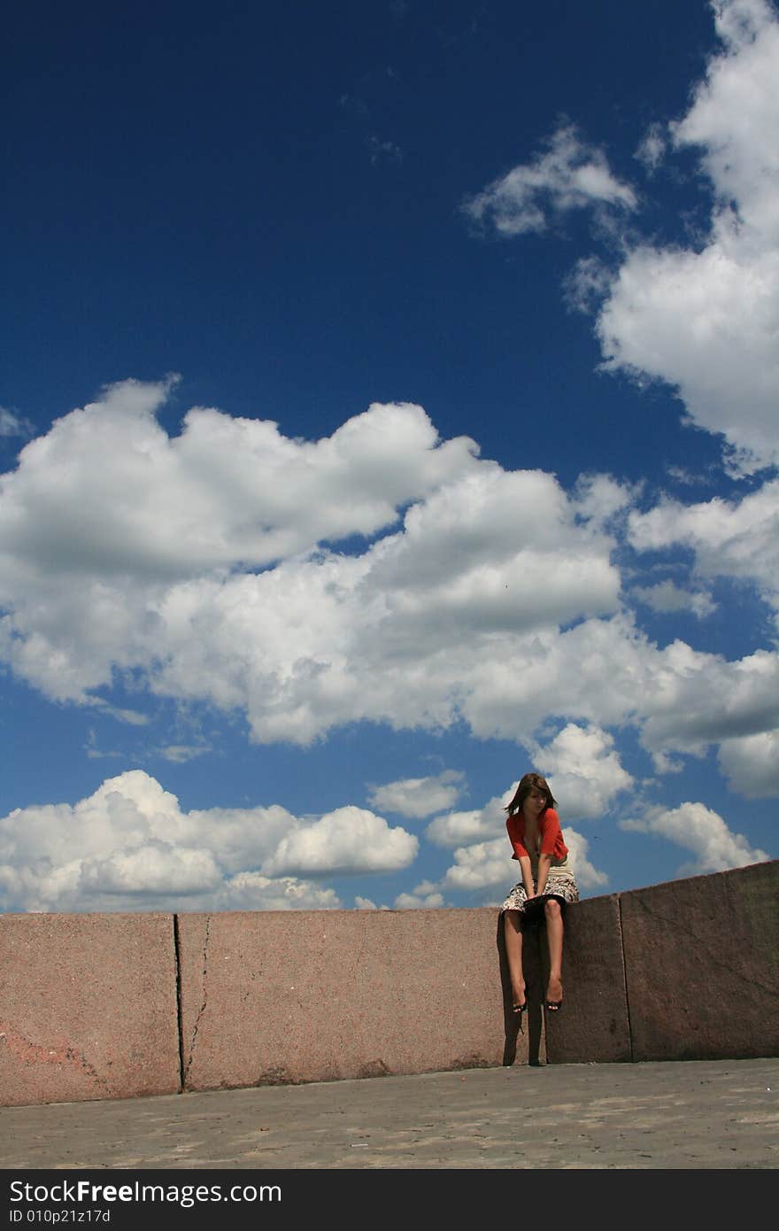 The young girl sits on a border of a roof on a background of the is bright-dark blue sky with snow-white clouds. The young girl sits on a border of a roof on a background of the is bright-dark blue sky with snow-white clouds