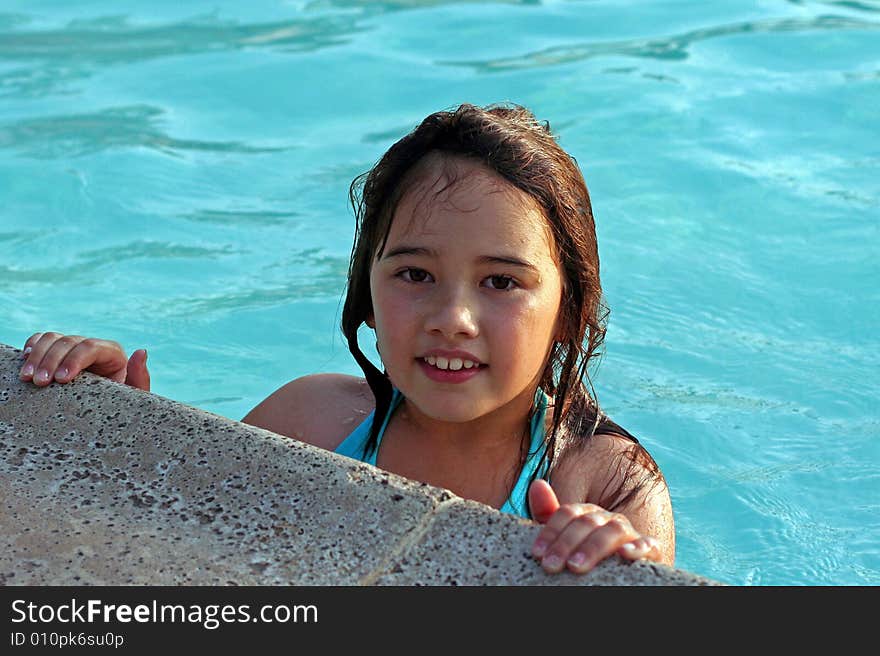 A smiling 6-year-old girl holds onto the edge of a swimming pool. She has big brown eyes and brown hair and is of Asian and Caucasian background, multi-racial. A smiling 6-year-old girl holds onto the edge of a swimming pool. She has big brown eyes and brown hair and is of Asian and Caucasian background, multi-racial.