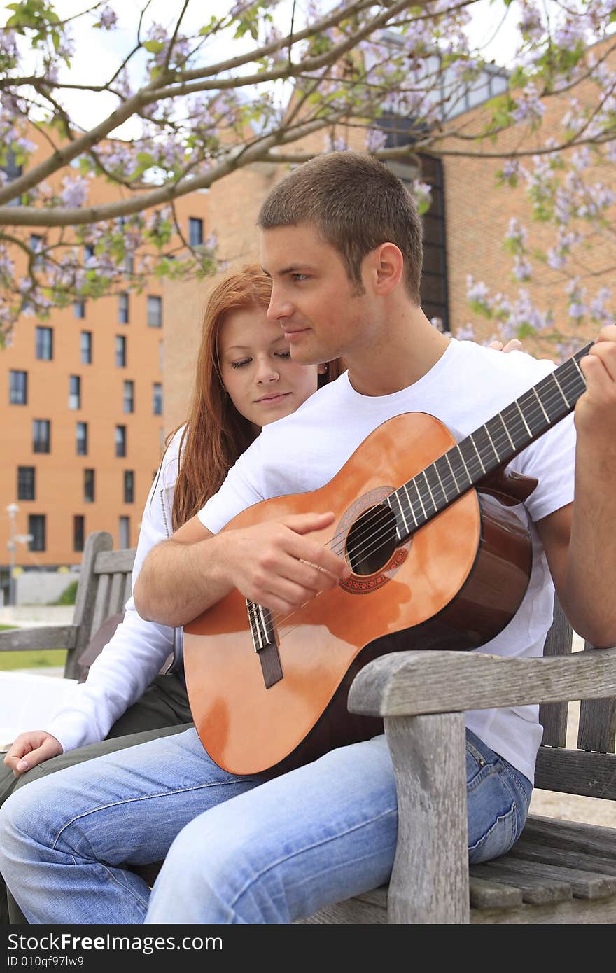 Young man plays for young girl on his guitar, it listens to him, both sits on a bank in the park. Young man plays for young girl on his guitar, it listens to him, both sits on a bank in the park