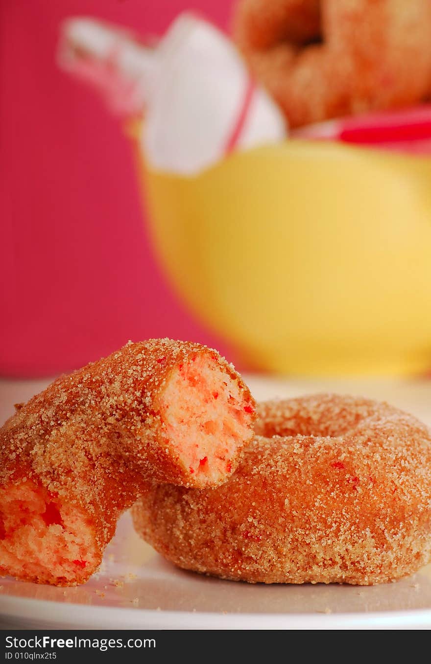 Two freshly made strawberry doughnuts with a bowl of doughnuts in the background. Two freshly made strawberry doughnuts with a bowl of doughnuts in the background