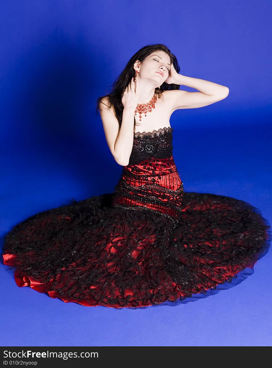 Beautiful young woman wearing a red evening gown sitting in front of a blue background