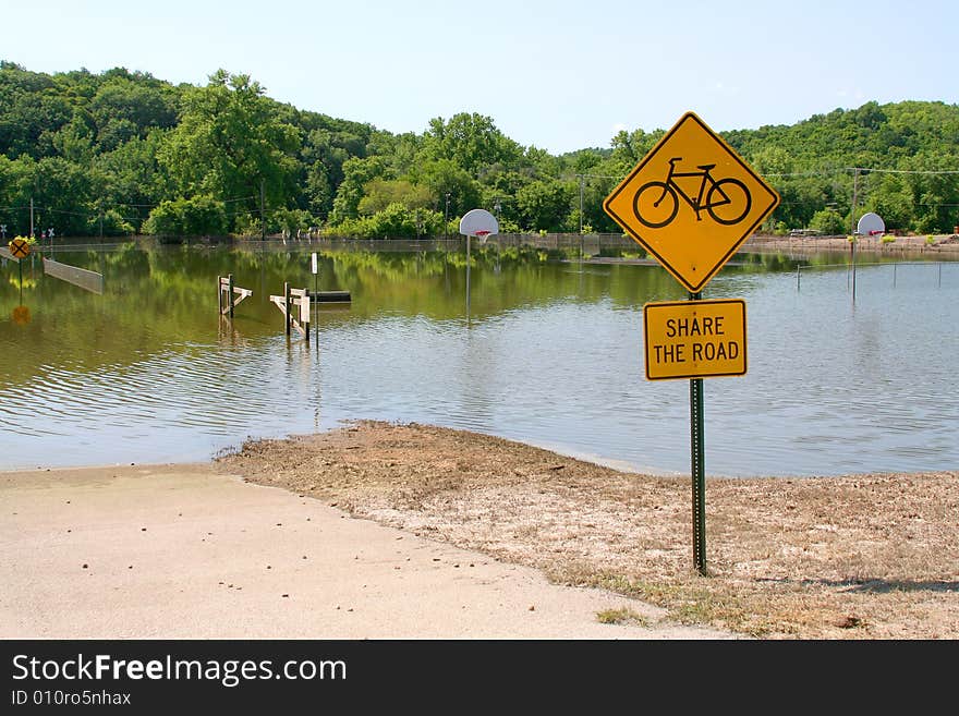 Flood waters share the road with a bicycle sign. A basketball court in the background is likewise submerged. Flood waters share the road with a bicycle sign. A basketball court in the background is likewise submerged.