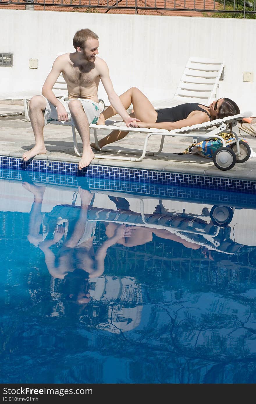 A man and a woman are relaxing next to a pool. The man is sitting up and looking at the woman. The woman is laying down and looking at the man. Vertically framed photo. A man and a woman are relaxing next to a pool. The man is sitting up and looking at the woman. The woman is laying down and looking at the man. Vertically framed photo.