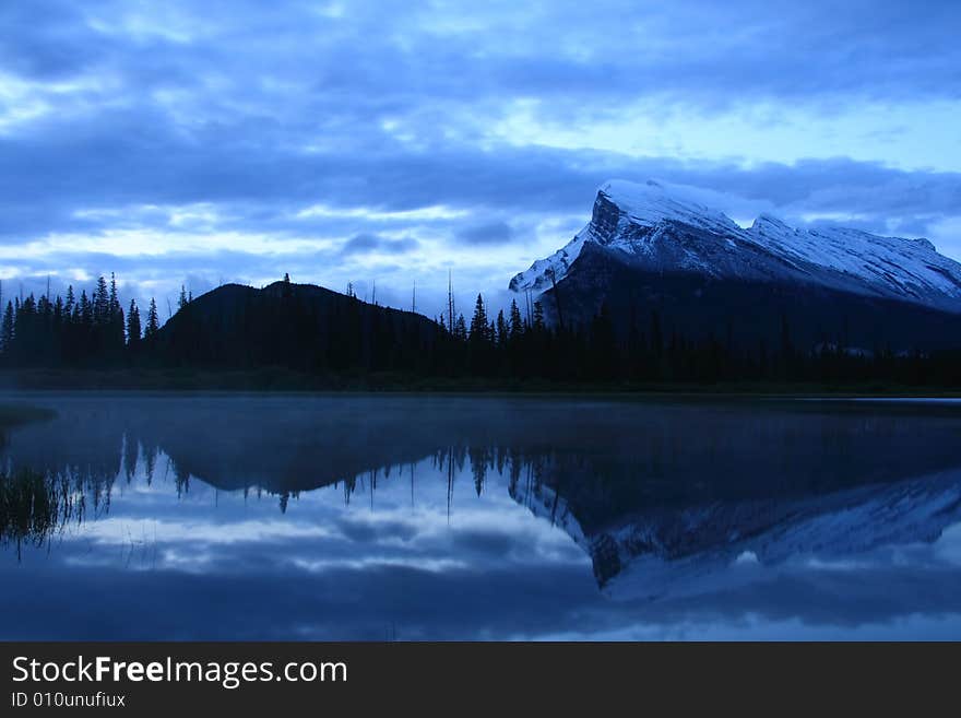 Mountains and forest reflected in a calm lake as the morning fills the sky with light. Mountains and forest reflected in a calm lake as the morning fills the sky with light