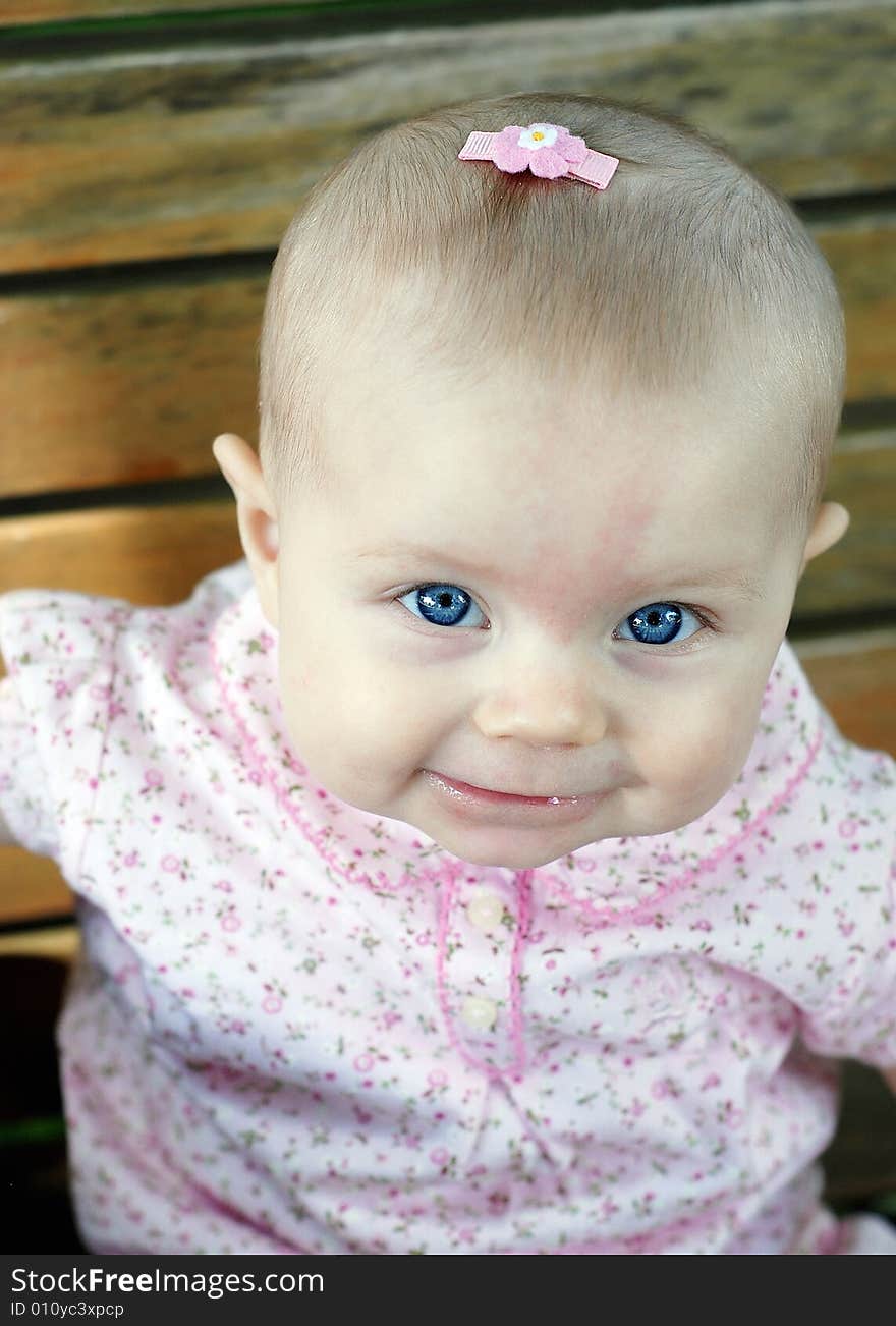 A young baby, wearing a pink bow in her hair and pink clothes, sits on a wooden park bench, smiling. Vertically framed shot. A young baby, wearing a pink bow in her hair and pink clothes, sits on a wooden park bench, smiling. Vertically framed shot.