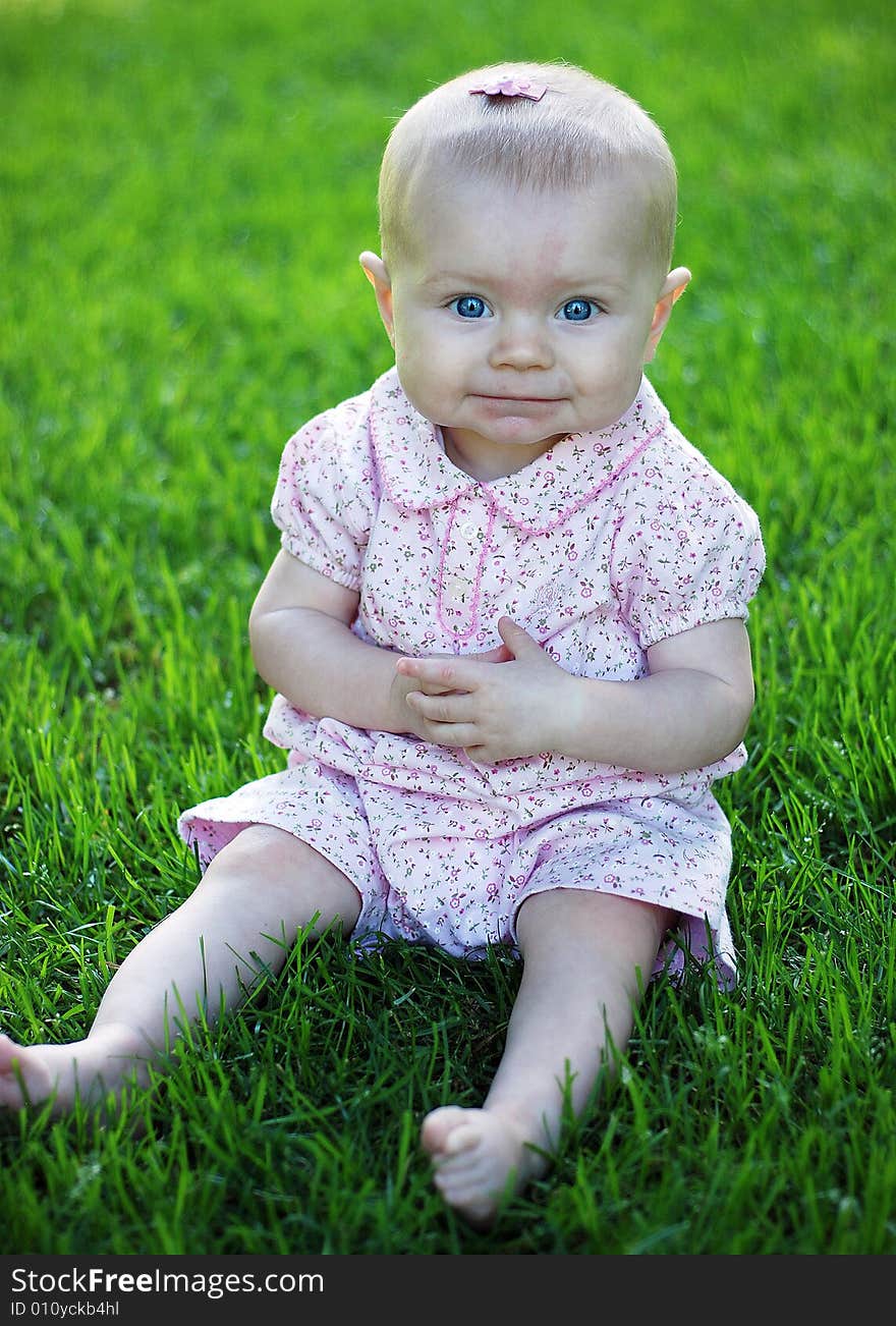 A young baby, wearing pink, sits in a grassy field, smiling. Vertically framed shot. A young baby, wearing pink, sits in a grassy field, smiling. Vertically framed shot.