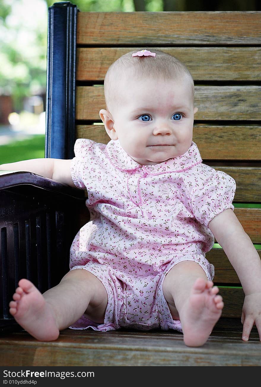 A young baby smiling while looking away, sits on a wooden park bench, wearing pink clothes. Vertically framed shot. A young baby smiling while looking away, sits on a wooden park bench, wearing pink clothes. Vertically framed shot.