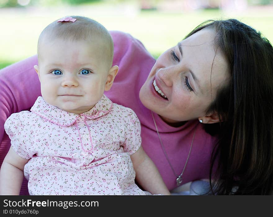 A mother, lying down, next to her baby, dressed in pink, sitting. Both, mother and baby, are smiling. Horizontally framed shot. A mother, lying down, next to her baby, dressed in pink, sitting. Both, mother and baby, are smiling. Horizontally framed shot.