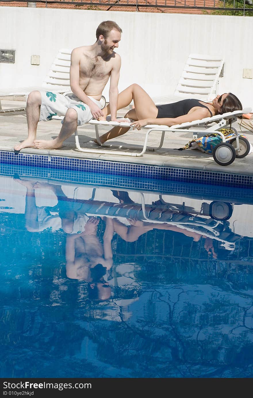 A young couple, sharing a poolside reclining chair, the woman lies down, as the man sits by her feet. - vertically framed. A young couple, sharing a poolside reclining chair, the woman lies down, as the man sits by her feet. - vertically framed