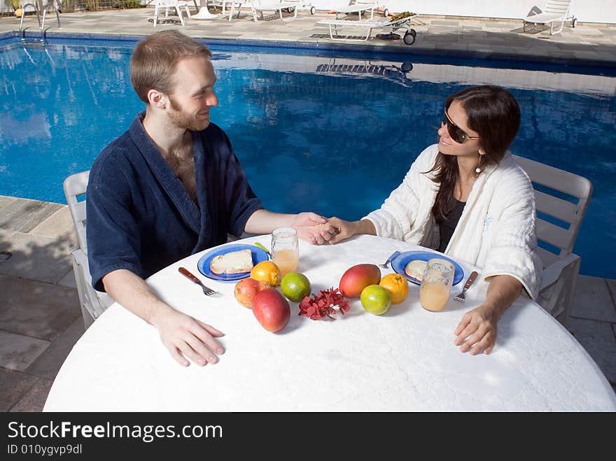 A couple, sitting at a table with fruits on it, near the pool, stare and smile, while holding hands. - horizontally framed. A couple, sitting at a table with fruits on it, near the pool, stare and smile, while holding hands. - horizontally framed