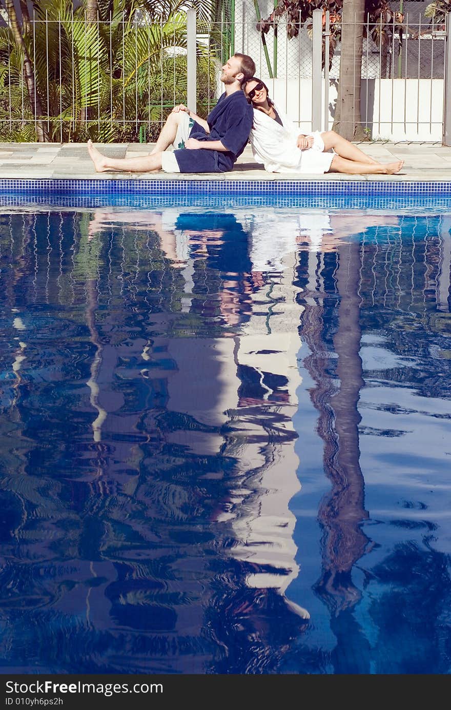 A man and a woman are relaxing together beside a pool.  They are leaning against each other.  The man is looking away from the camera and the woman is looking at it.  Vertically framed photo. A man and a woman are relaxing together beside a pool.  They are leaning against each other.  The man is looking away from the camera and the woman is looking at it.  Vertically framed photo.