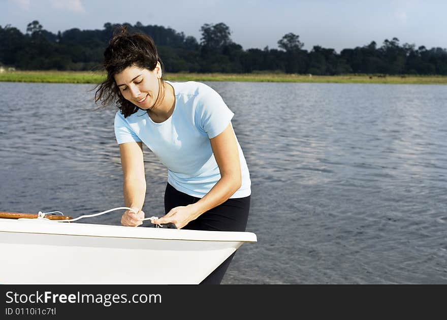 A woman is standing next to a sailboat on the water.  She is tying a knot in a rope, smiling, and looking away from the camera.  Horizontally framed shot. A woman is standing next to a sailboat on the water.  She is tying a knot in a rope, smiling, and looking away from the camera.  Horizontally framed shot.