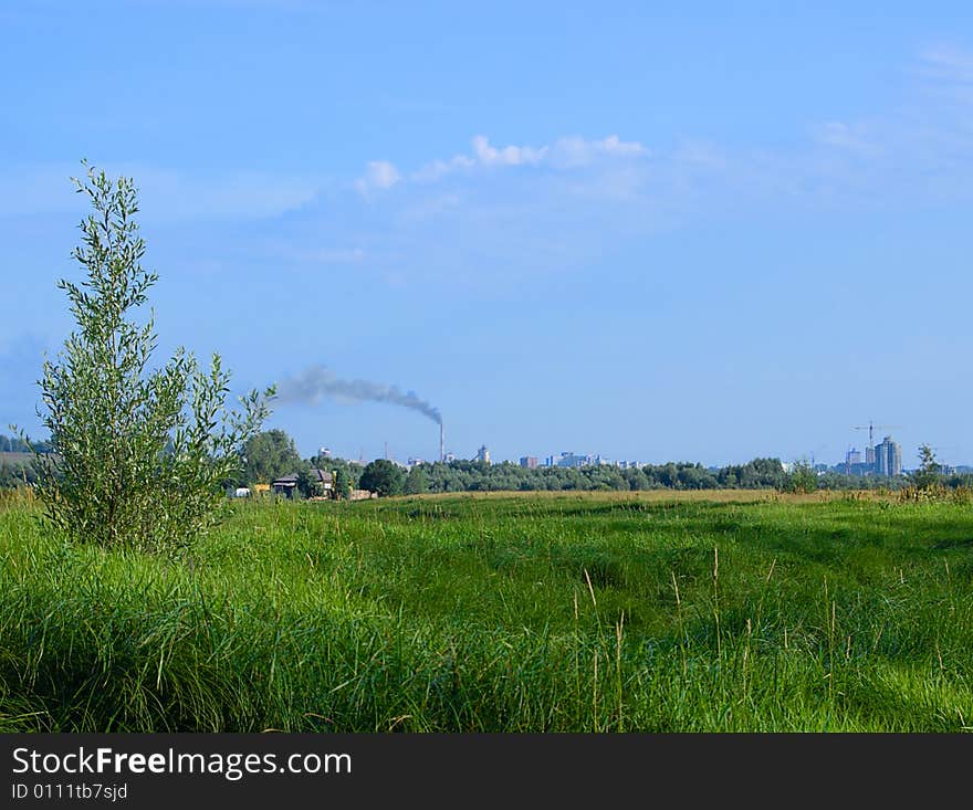 Smoking stack and  building in the background, green grass and tree in the foreground. Smoking stack and  building in the background, green grass and tree in the foreground