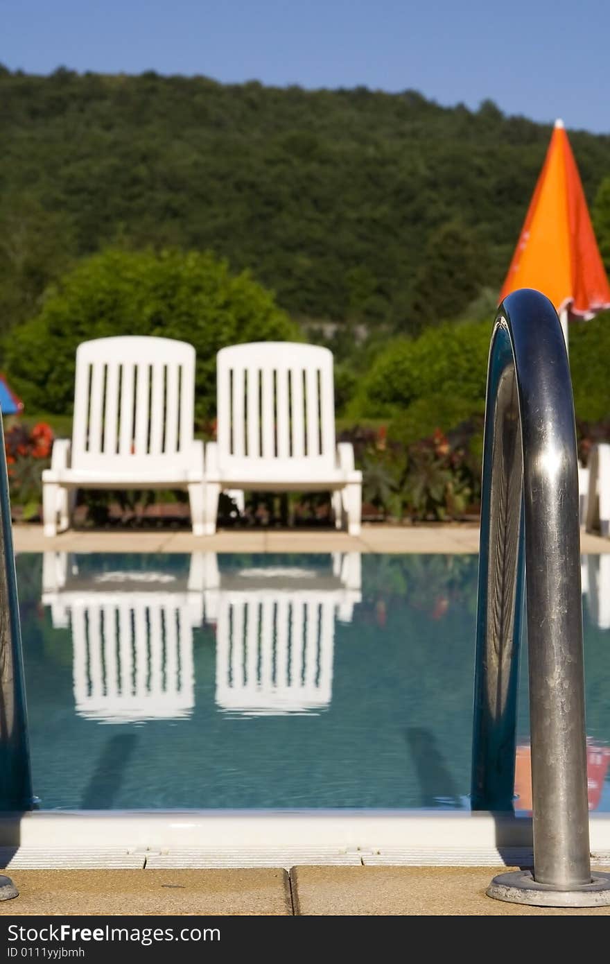 Two white sunloungers reflected in a blue swimming pool, next to an orange umbrella.  The focus is on the metal handrail in the foreground. Two white sunloungers reflected in a blue swimming pool, next to an orange umbrella.  The focus is on the metal handrail in the foreground.