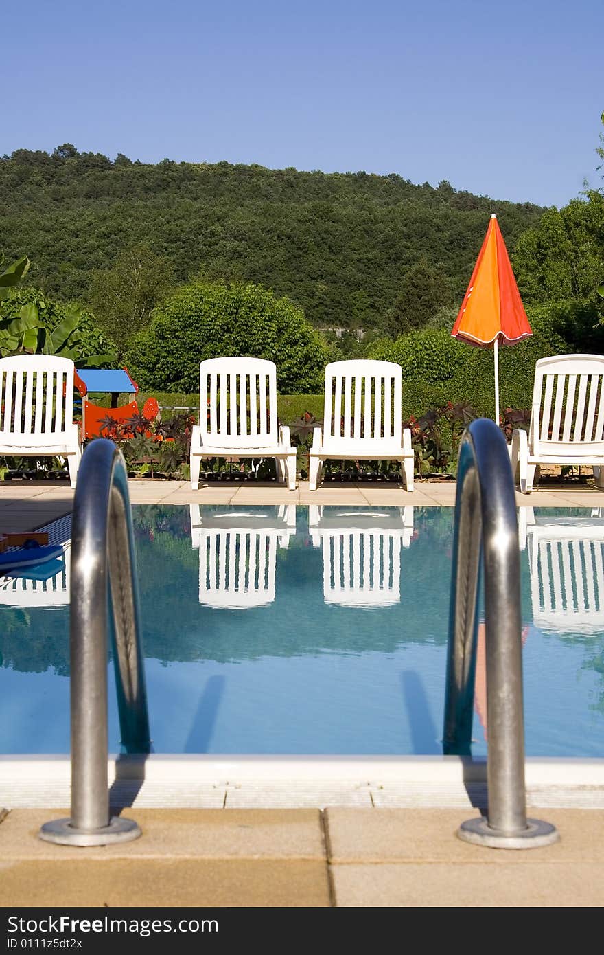 White sunloungers reflected in a blue swimming pool, next to an orange umbrella. White sunloungers reflected in a blue swimming pool, next to an orange umbrella