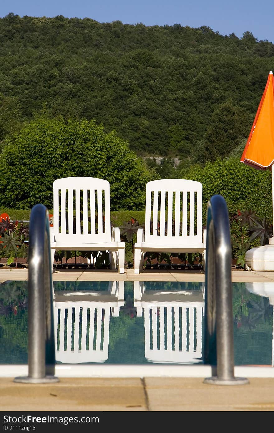 Two white sunloungers reflected in a blue swimming pool, next to an orange umbrella. Two white sunloungers reflected in a blue swimming pool, next to an orange umbrella