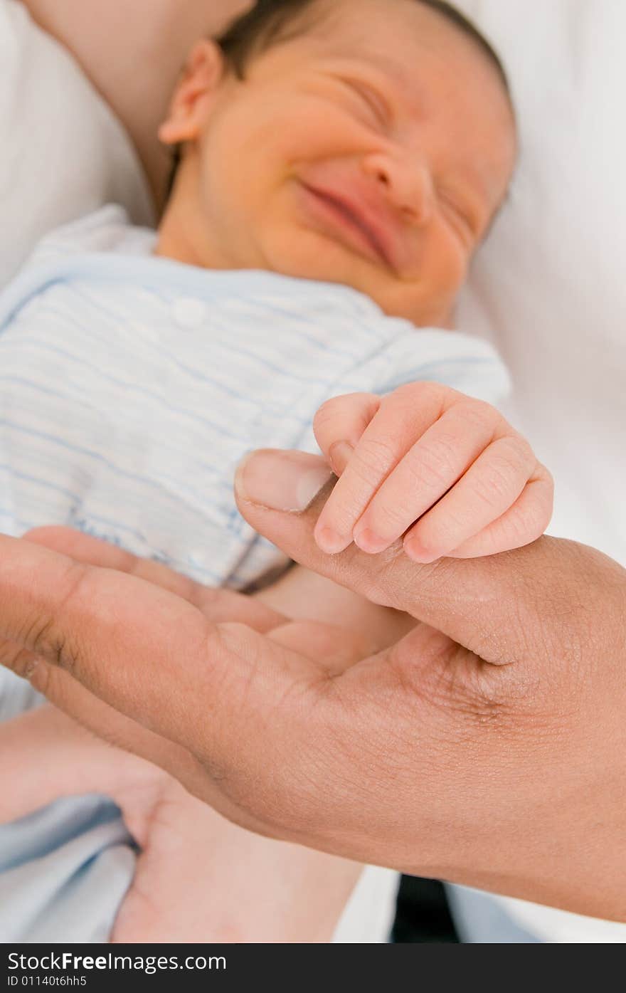 Baby's hand holding father's thumb with baby's grinning face in background. Baby's hand holding father's thumb with baby's grinning face in background