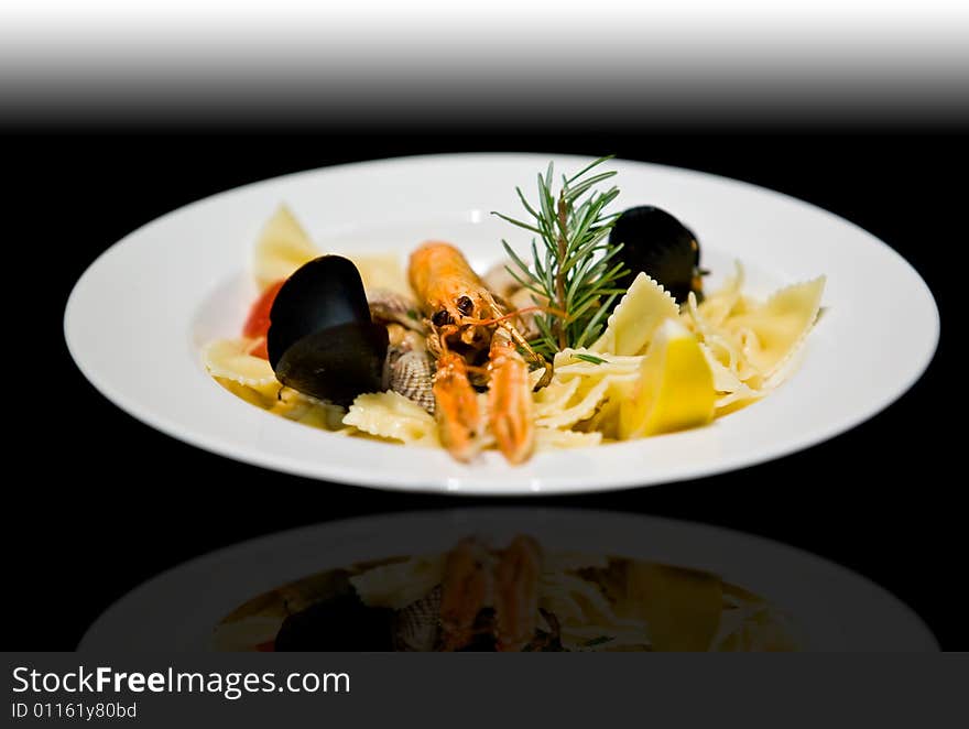 Langoustine, clam, and mussel pasta appetizer served with rosemary and lemon in a white plate over a graduate black background. Langoustine, clam, and mussel pasta appetizer served with rosemary and lemon in a white plate over a graduate black background.