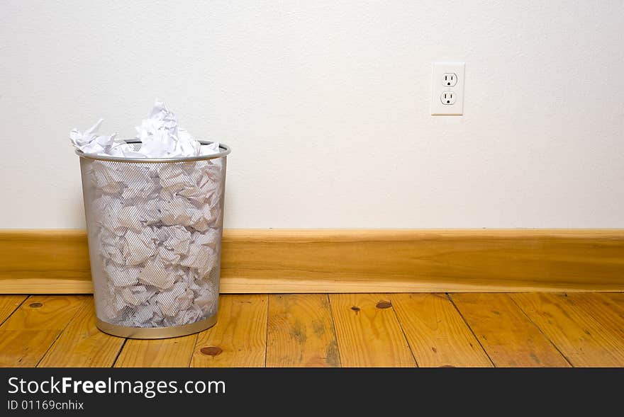 A wire mesh office trash can with crumpled paper on a wooden floor with a white wall, includes copy space. A wire mesh office trash can with crumpled paper on a wooden floor with a white wall, includes copy space