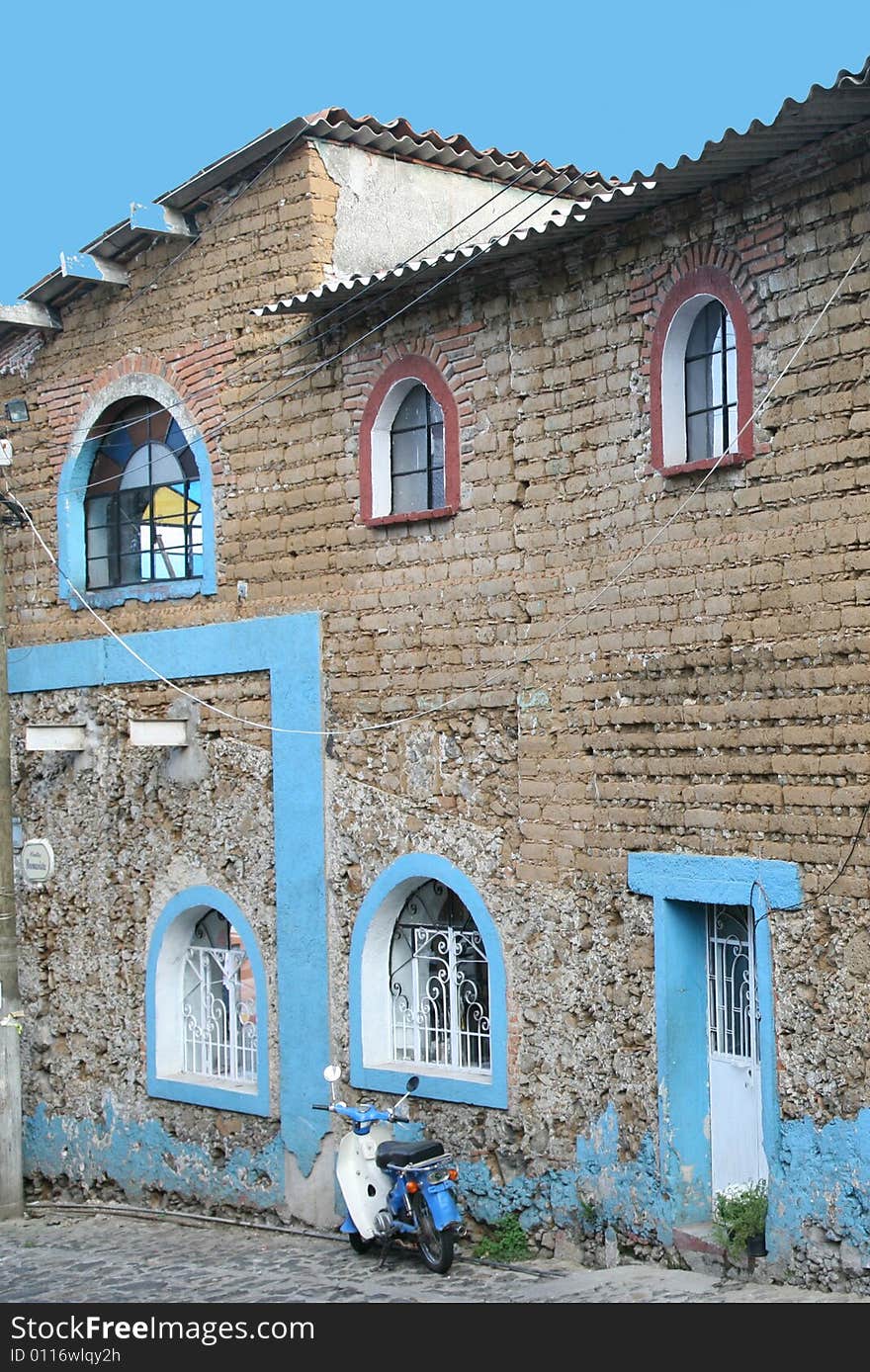 Blue motorbike parked alongside a stone building accented in blue trim, image of a poor Mexican village.