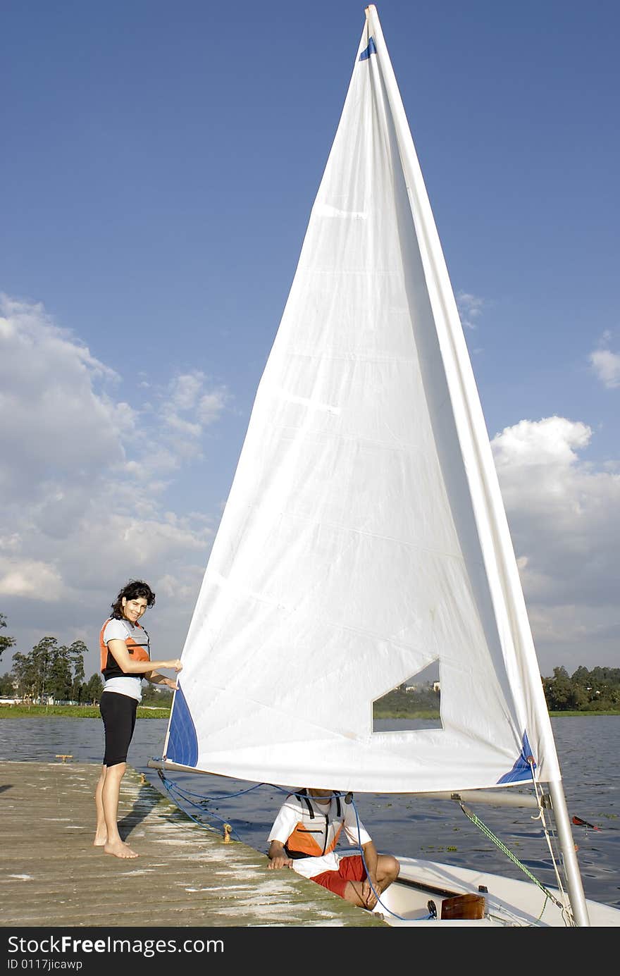 A woman and man are next to a sailboat.  The man is sitting inside the boat.  The woman is standing beside it fixing the sail and smiling at the camera.  Vertically framed shot. A woman and man are next to a sailboat.  The man is sitting inside the boat.  The woman is standing beside it fixing the sail and smiling at the camera.  Vertically framed shot.