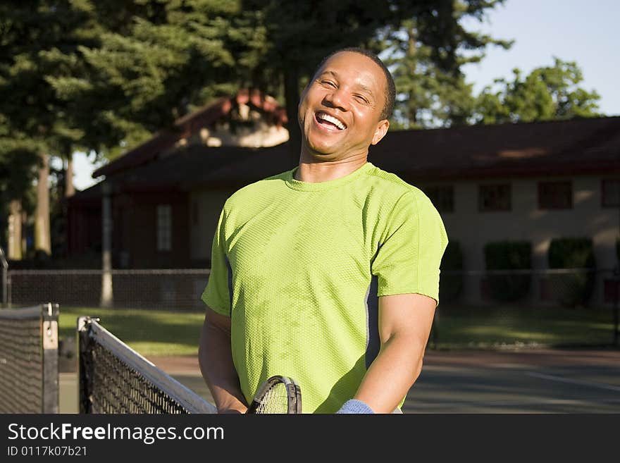 A man is outside on a tennis court.  He is leaning against the net and smiling at the camera. A man is outside on a tennis court.  He is leaning against the net and smiling at the camera.