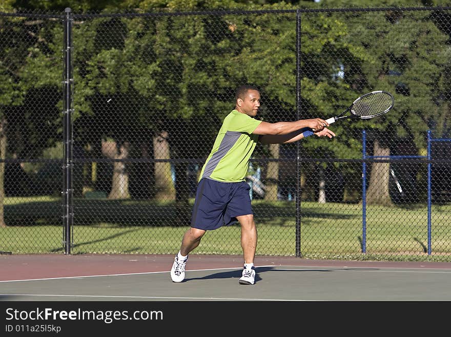 A man is outside on a tennis court playing tennis. He is looking at the tennis ball after he just hit it with his racket. A man is outside on a tennis court playing tennis. He is looking at the tennis ball after he just hit it with his racket.