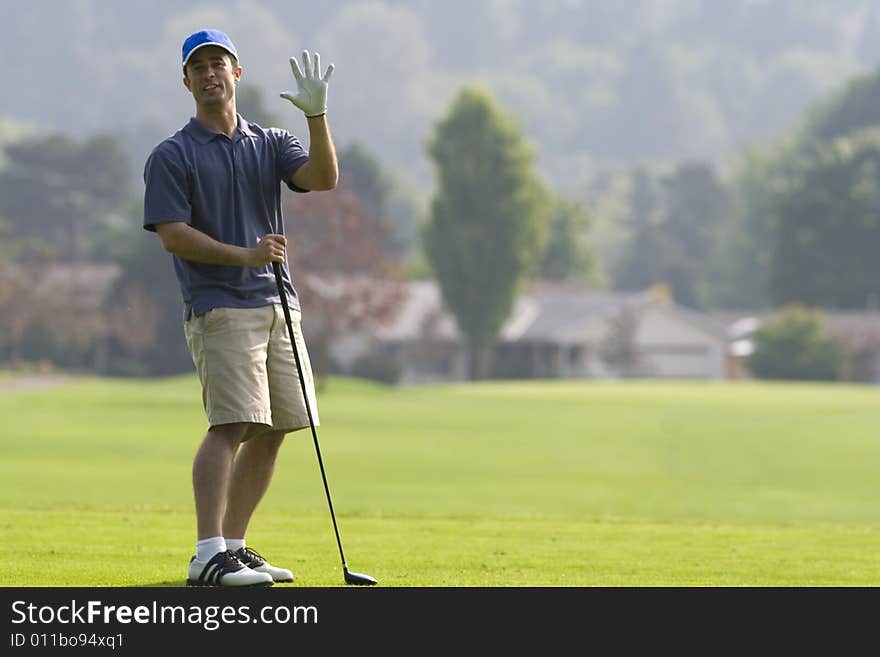 Man standing on golf course waving his right hand at the camera with left hand resting on golf club - horizontally framed photo. Man standing on golf course waving his right hand at the camera with left hand resting on golf club - horizontally framed photo