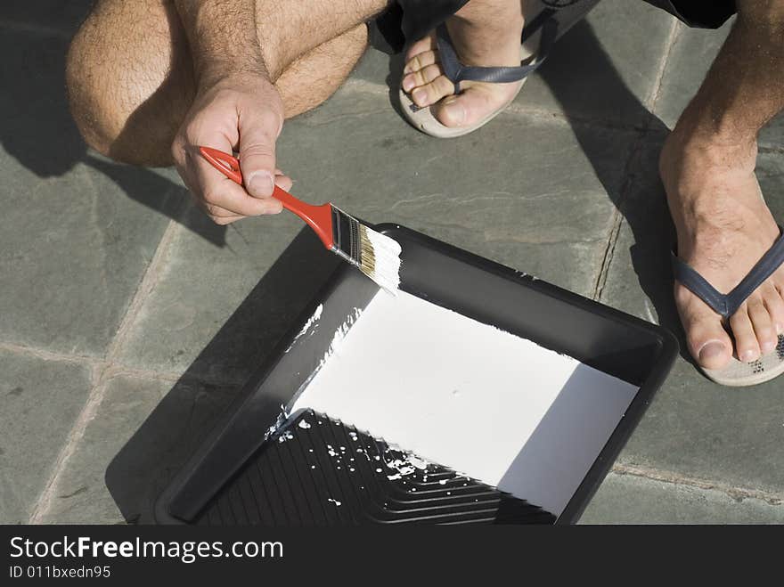 A man, kneels, dipping his paintbrush into a platter of white paint. Horizontally framed shot. A man, kneels, dipping his paintbrush into a platter of white paint. Horizontally framed shot.