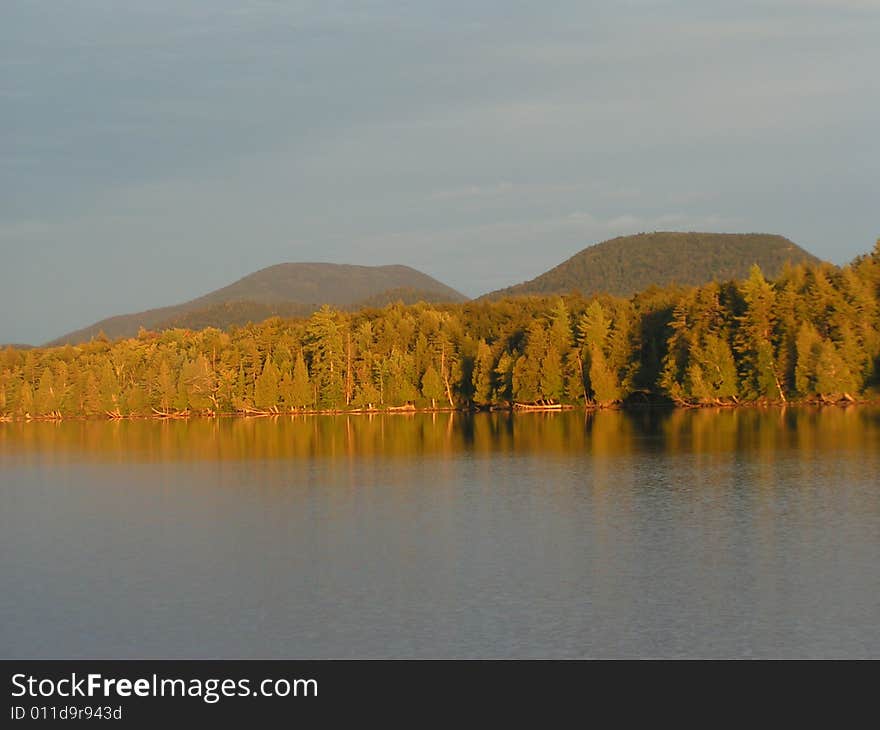 Golden sunlight at sunset on trees and twin mountains behind lake in Adirondacks, New York. Golden sunlight at sunset on trees and twin mountains behind lake in Adirondacks, New York.