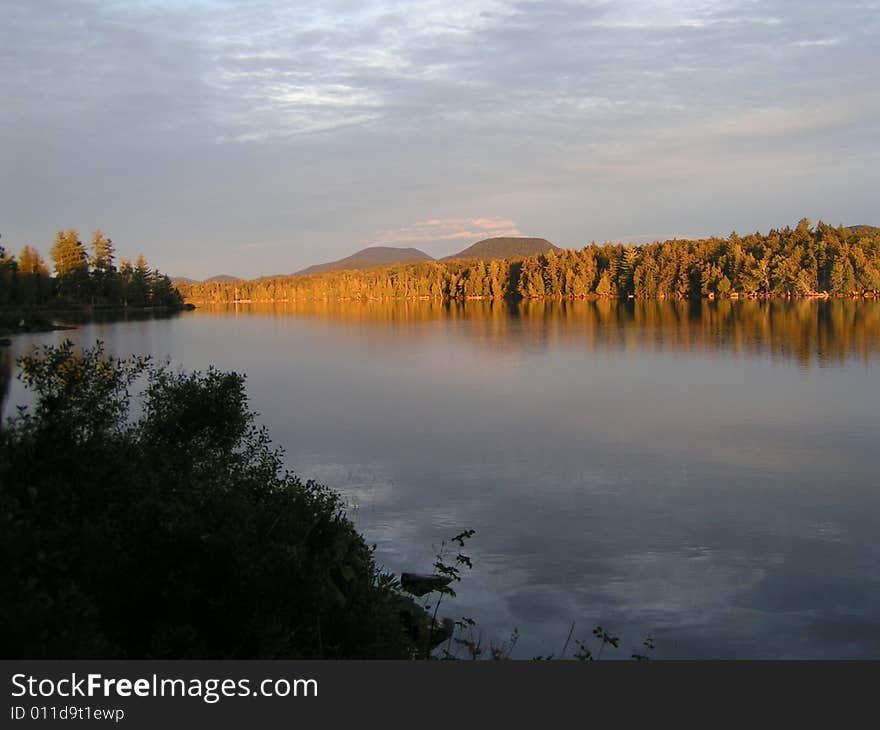 Golden sunlight at sunset on trees and twin mountains behind lake in Adirondacks, New York. Golden sunlight at sunset on trees and twin mountains behind lake in Adirondacks, New York.