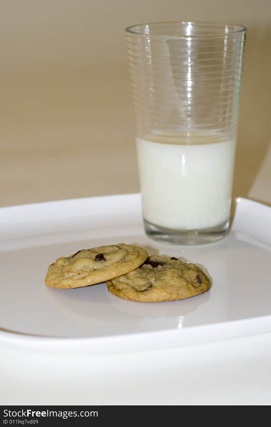 A pair of chocolate chip cookies sits stacked on a white surface, with a glass of milk in the background. A pair of chocolate chip cookies sits stacked on a white surface, with a glass of milk in the background.