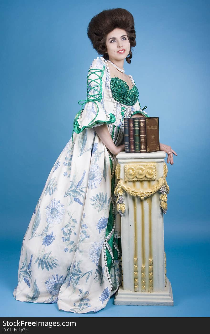 Beautiful 18th century elegant lady wearing vintage fashion dress and posing in front of a baroque column with books. Beautiful 18th century elegant lady wearing vintage fashion dress and posing in front of a baroque column with books.