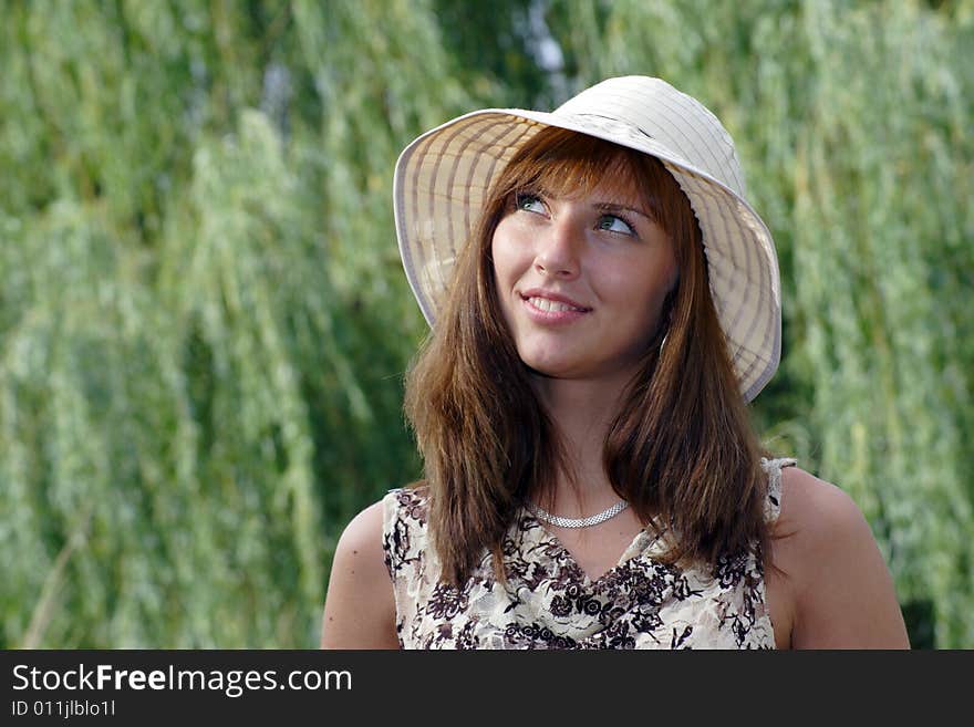 Beautiful smiling woman in the hat looks somewhere upward. A green tree branches on the smear background.