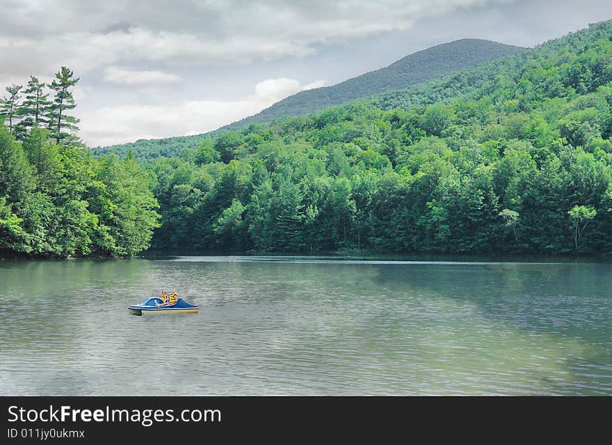 One of Vermonts wonderful lakes shows the shimmering color of emerald and the reflections of trees and clouds on the water as 2 children are enjoying the use of a a paddle boat on  the water. One of Vermonts wonderful lakes shows the shimmering color of emerald and the reflections of trees and clouds on the water as 2 children are enjoying the use of a a paddle boat on  the water