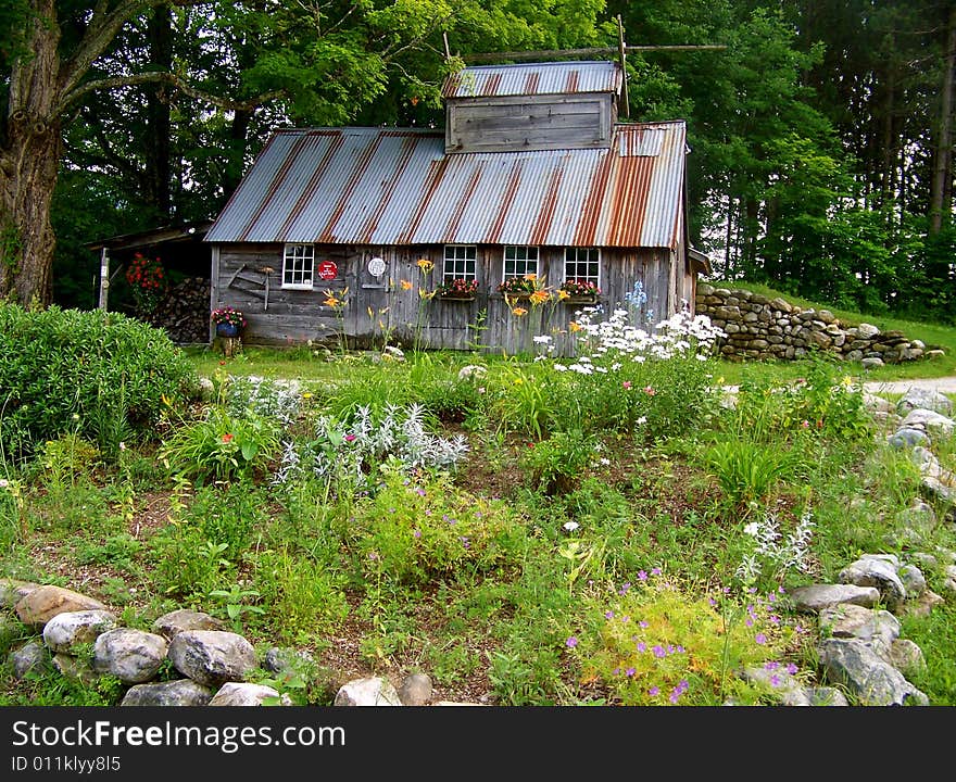 A beautiful sugar shack adorned with flowers in the window boxes for summer sits on a little hill surrounded with stone walls.