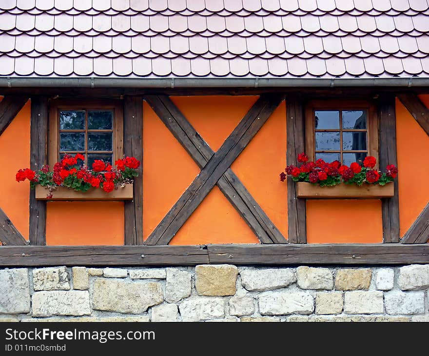 The old fashioned country home detail. Two wooden windows with red flowers in flower-pots. Post and pan, stone wall and new roof. The old fashioned country home detail. Two wooden windows with red flowers in flower-pots. Post and pan, stone wall and new roof.