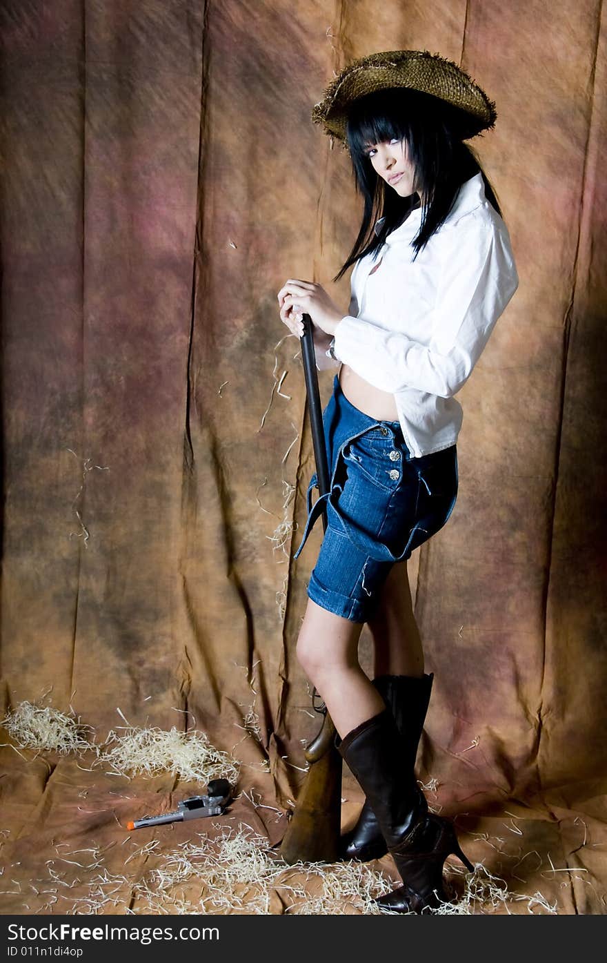 Portrait of a young woman with black hair wearing a white shirt, straw hat white shirt and folded down denim dungarees and pink lipstick. She is holding a shotgun. Portrait of a young woman with black hair wearing a white shirt, straw hat white shirt and folded down denim dungarees and pink lipstick. She is holding a shotgun.