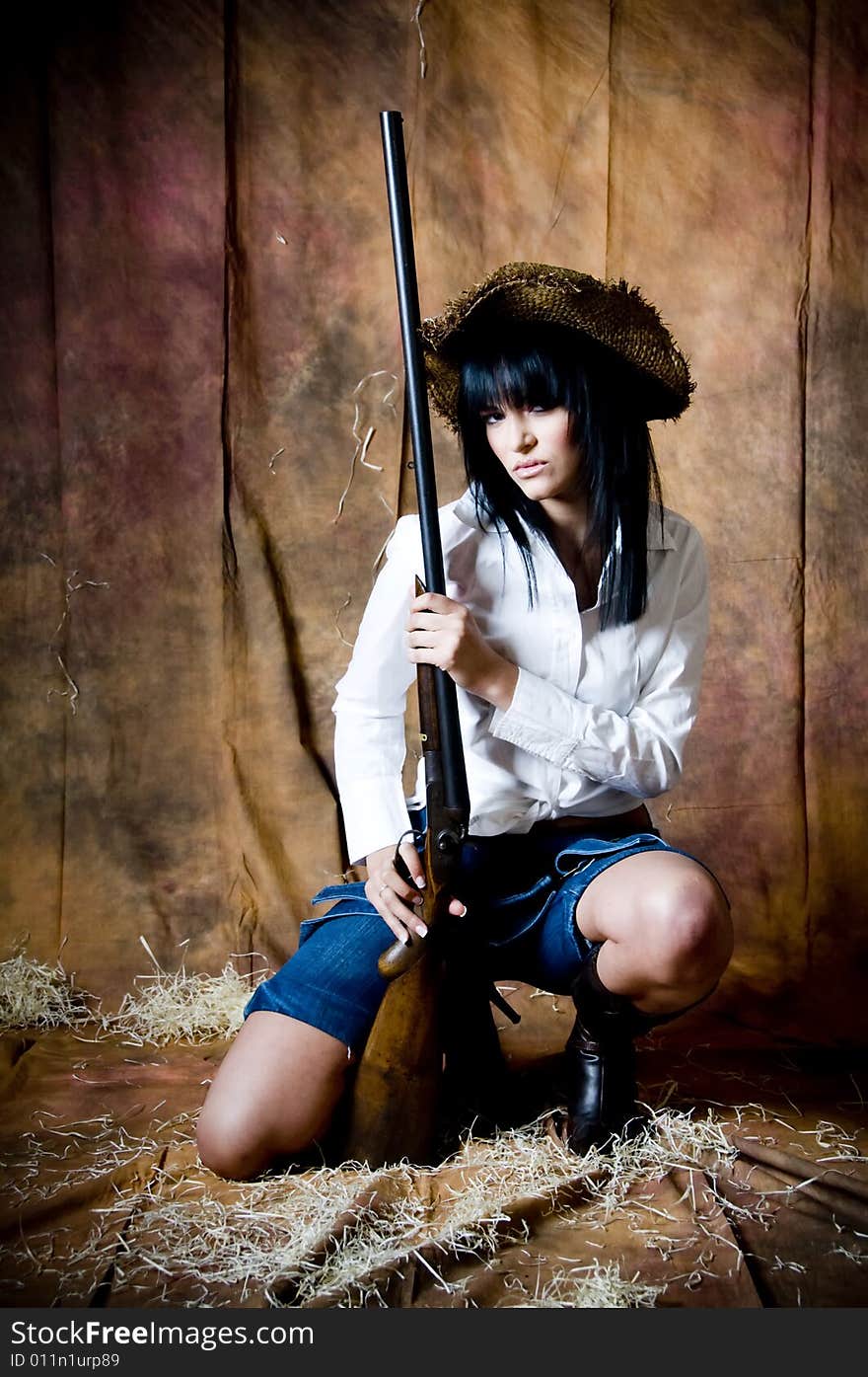 A young woman kneels holding an old shotgun. She wears a straw hat, white shirt and denim dungarees folded down. A young woman kneels holding an old shotgun. She wears a straw hat, white shirt and denim dungarees folded down.