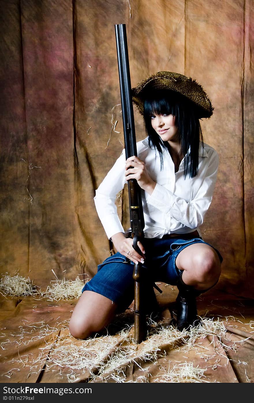 A young smiling woman kneels holding an old shotgun. She wears a straw hat, white shirt and denim dungarees folded down. A young smiling woman kneels holding an old shotgun. She wears a straw hat, white shirt and denim dungarees folded down.