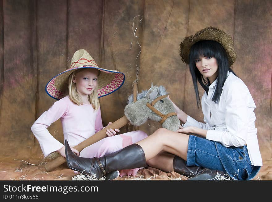 A landscape of a young woman in denim and white, and a young girl in a pink dress sit together wearing straw hats and playing with a toy horse. A landscape of a young woman in denim and white, and a young girl in a pink dress sit together wearing straw hats and playing with a toy horse.