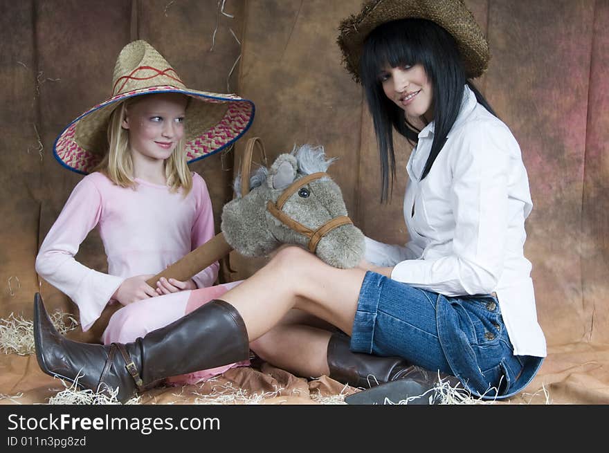 A landscape of a young woman in denim and white, and a young girl in a pink dress - both smiling - sit together wearing straw hats and playing with a toy horse. A landscape of a young woman in denim and white, and a young girl in a pink dress - both smiling - sit together wearing straw hats and playing with a toy horse.