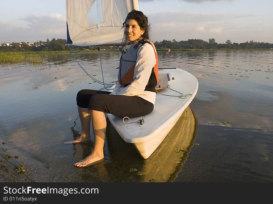 Woman sitting on bow of sailboat. Feet resting on ground in shallow water. Looking at camera and smiling. Horizontally framed photo. Woman sitting on bow of sailboat. Feet resting on ground in shallow water. Looking at camera and smiling. Horizontally framed photo.