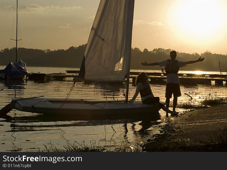 Woman sitting on sailboat. Man standing on shore in front of sunset. Horizontally framed photo. Woman sitting on sailboat. Man standing on shore in front of sunset. Horizontally framed photo