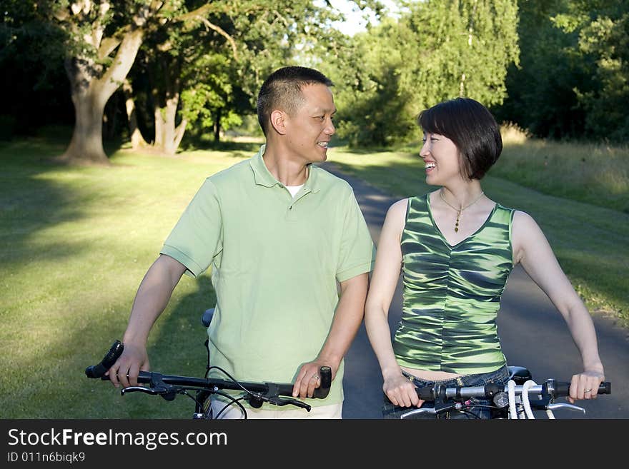 Asian couple standing alongside bicycles smiling at eachother. Couple standing in park. Horizontally framed photo. Asian couple standing alongside bicycles smiling at eachother. Couple standing in park. Horizontally framed photo.