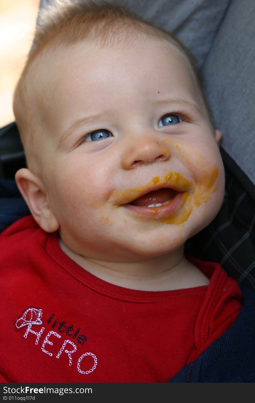 Newborn baby sitting and smiling with food on face.  The child has a red shirt and blue eyes. Newborn baby sitting and smiling with food on face.  The child has a red shirt and blue eyes