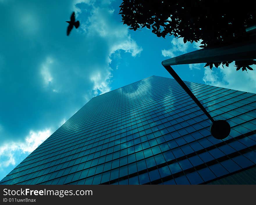 This is an image of a building. The dominant color is blue. This image conveys the idea of reaching for success. There are multiple objects that project height or rising up. A tree, a street light, a bird, the building, clouds,and sky at different heights shows there is always something higher to be acquired. This is an image of a building. The dominant color is blue. This image conveys the idea of reaching for success. There are multiple objects that project height or rising up. A tree, a street light, a bird, the building, clouds,and sky at different heights shows there is always something higher to be acquired.