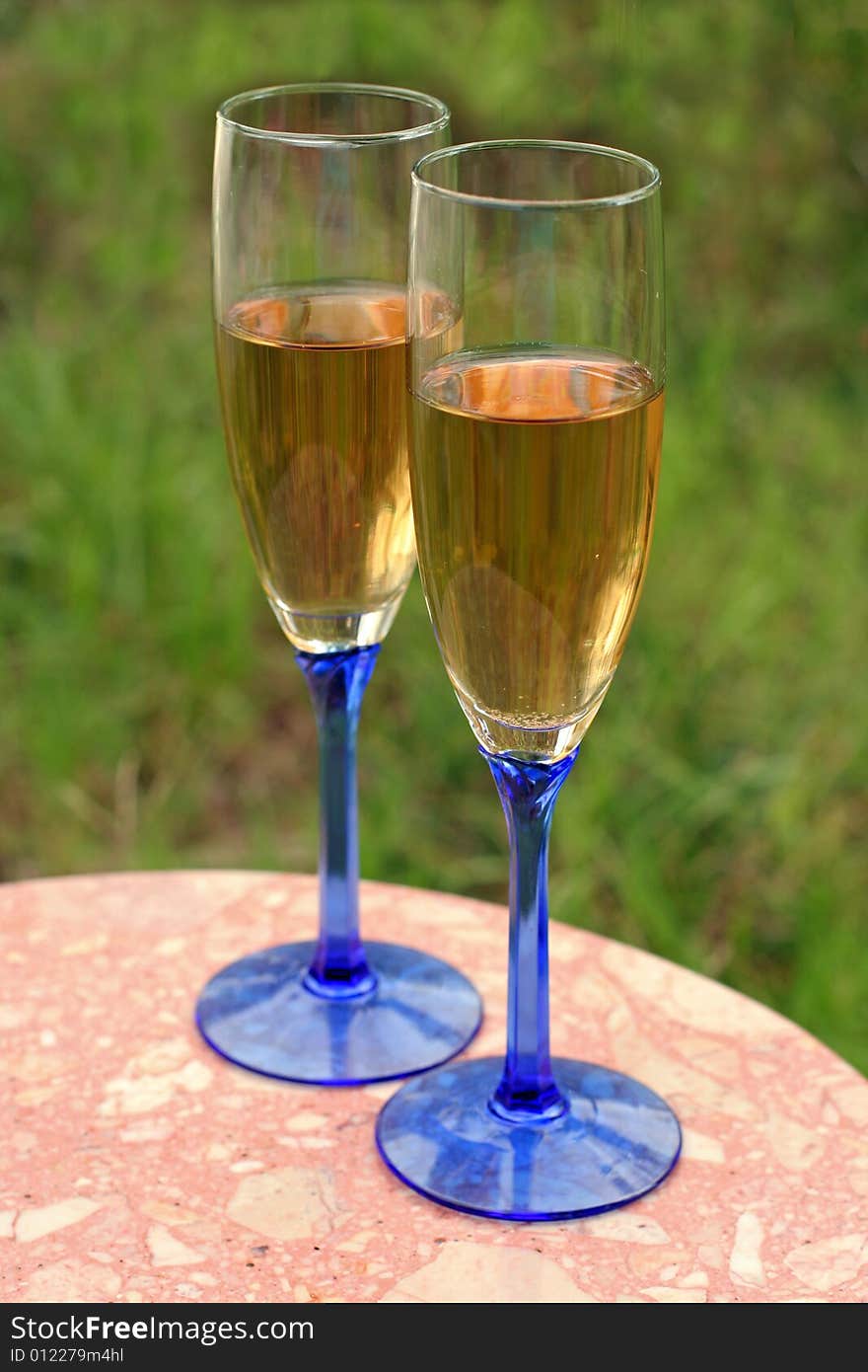 Two glasses of white wine in blue-stemmed glasses, on a small table with a grassy background. Two glasses of white wine in blue-stemmed glasses, on a small table with a grassy background.