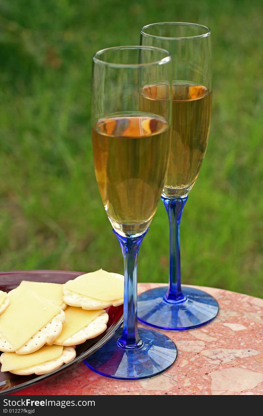 Two glasses of white wine in blue-stemmed glasses on a small marble table outside with a grassy background. Drinks are served with cheese and crackers on a red plate. Two glasses of white wine in blue-stemmed glasses on a small marble table outside with a grassy background. Drinks are served with cheese and crackers on a red plate.